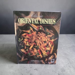 May include: A cookbook titled "Classic Oriental Dishes" stands upright. The cover features a vibrant image of a stir-fry dish with vegetables and peanuts. The text "Classic Oriental Dishes" is displayed prominently at the top.