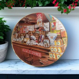 May include: A vintage-style ceramic plate featuring a colorful illustration of a grocery store interior with the text "I.G.A. STORES". The plate depicts a bustling scene with shelves stocked with various goods, including canned goods, a scale, and a cash register.