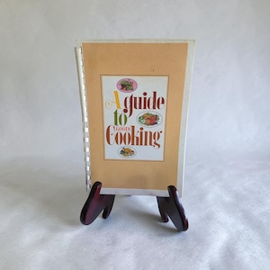 May include: A spiral-bound cookbook with a light tan cover featuring the text A guide to Good Cooking in brown and green lettering. The cover includes three small circular illustrations of food items, including a salad, a lobster, and a roasted chicken.