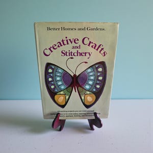 May include: A hardcover book titled Better Homes and Gardens Creative Crafts and Stitchery with a large, colorful butterfly illustration on the front cover. The butterfly features shades of purple, blue, green, and gold. The book rests on a stand.