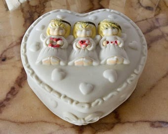 Puede incluir: Caja de corazón blanca con tres querubines que sostienen corazones rojos. Los querubines tienen el pelo rubio y llevan túnicas blancas con alas.