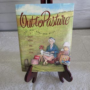 May include: A hardcover book titled Out to Pasture (but not over the hill) A Short Novel by Effie Leland Wilder. The cover features an illustration of an older person typing on a typewriter in a grassy field with a lamp, flowers, and books.