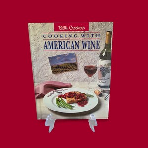 May include: A cookbook titled "Cooking with American Wine" by Betty Crocker. The cover features a photo of a meal with wine, a wine bottle, and a glass of red wine. The background is a textured, off-white surface.