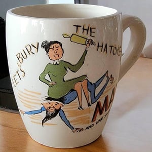 May include: A white ceramic mug with a cartoon illustration of a woman holding a rolling pin and a man with a beard. The text on the mug reads "Let's Bury The Hatchet" and "Make Mine A Have You".