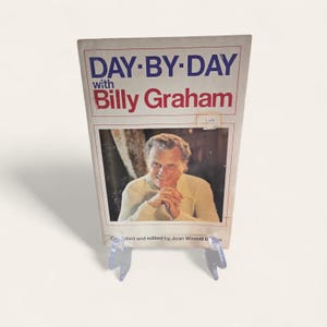May include: A vintage book titled "DAY-BY-DAY with Billy Graham" is displayed on a clear plastic stand. The book cover features a portrait of Billy Graham and text in red and blue. The book is compiled and edited by Joan Winmill Brown.