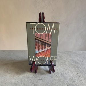 以下が含まれることがあります： トム・ウルフ著『虚栄の篝火』のハードカバー本。グレーの表紙の中央には、模様入りの絨毯の上にある面に反射した都市のスカイラインのイラストが描かれており、濃い茶色の木製スタンドに立てかけられている。