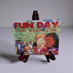 May include: A children's book titled "FUN DAY in Mrs. Walker's Class" is displayed on a dark brown wooden stand. The book cover features illustrations of children and the author's name. The stand has a unique, decorative design.
