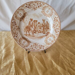 May include: A white ceramic plate with a brown and white design. The plate features a scene of soldiers on horseback and a building in the background. The text "Bataille de Montmirail et de Montereau 1814" is printed on the plate.