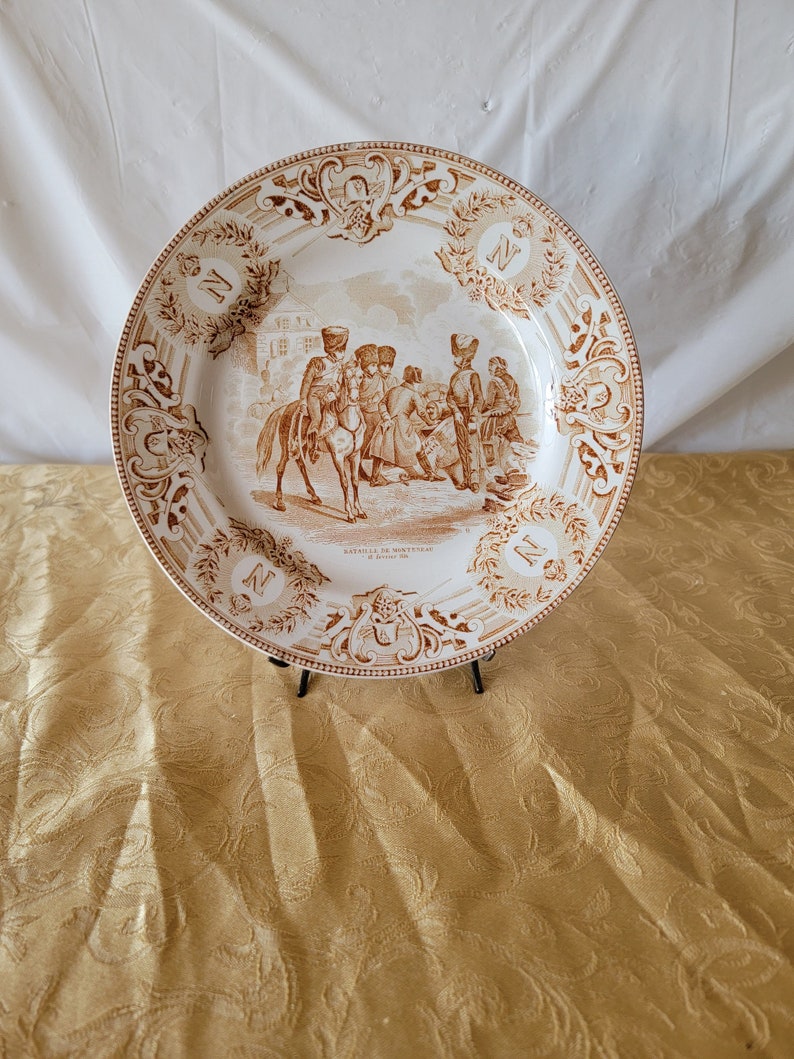 May include: A white ceramic plate with a brown and white design. The plate features a scene of soldiers on horseback and a building in the background. The text "Bataille de Montmirail et de Montereau 1814" is printed on the plate.