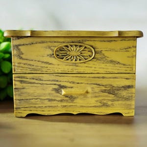 Pode incluir: Uma pequena caixa de madeira com duas gavetas e um acabamento de pintura amarela desgastada. A gaveta superior tem um desenho floral esculpido no centro.