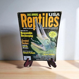 Peut inclure: Un magazine intitulé "2003 Annual Reptiles USA" est présenté sur un présentoir en bois rouge foncé. La couverture du magazine présente un reptile vert et beige avec le texte "Vital Info for Responsible Herpkeeping" et "Crested Geckos: Latest Color Morphs."