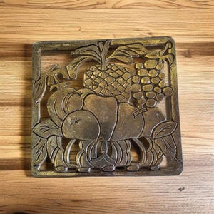May include: A square, metallic trivet with a raised relief design of various fruits, including a pineapple, grapes, and apples. The trivet has a gold and bronze color scheme and is set against a wooden background. The frame is square.