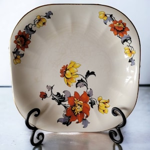 May include: A white ceramic square serving dish with a gold rim and a floral design in shades of yellow, orange, and red.