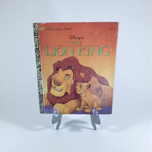 May include: A vintage copy of Disney's "The Lion King" Little Golden Book. The cover features Simba and Mufasa with a sunset background. The title is in green and the book is displayed on a clear plastic stand.