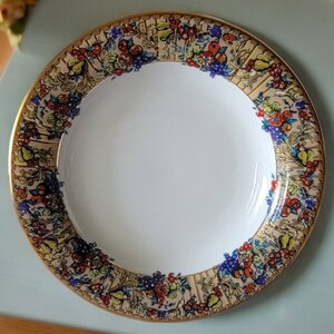 May include: A white ceramic plate with a gold rim and a colorful floral design featuring fruits and leaves. The design is around the edge of the plate.