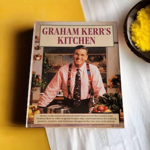 May include: A cookbook titled "Graham Kerr's Kitchen" features a photo of a man in a pink shirt and suspenders, smiling and looking at the camera. The book cover has a green and brown background with a variety of vegetables and herbs.