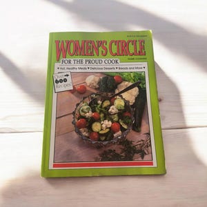 以下が含まれることがあります： 「Women's Circle For The Proud Cook」というタイトルの緑色の料理本。表紙にはボウルに入ったサラダの写真が載っている。表紙には「Over 600 Recipes!」という文字が書かれている。