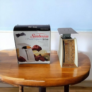 May include: A white Sunbeam 1 pound capacity diet scale with a black and white box showing fruit and a scale. The scale has a white dial with black numbers and a black Sunbeam logo.