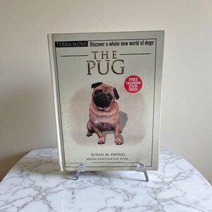 May include: A hardcover book titled "The Pug" featuring a seated pug on the cover. The book has the text "Discover a whole new world of dogs" and "Free Training DVD Inside". The book is displayed on a clear plastic stand.