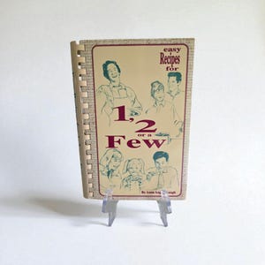 May include: A vintage cookbook titled "Easy Recipes for 1, 2 or a Few" with a spiral binding. The cover features illustrations of people and text in a retro style. The book is displayed on a clear plastic stand.