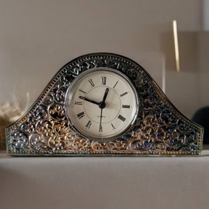 May include: A silver and gold decorative clock with an ornate design. The clock face has black Roman numeral hour markers and black hands.