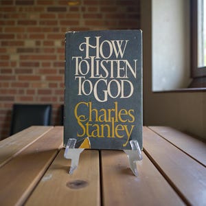 Peut inclure: Un livre intitulé "How to Listen to God" de Charles Stanley. La couverture est bleu foncé avec des lettres blanches et dorées. Le livre est présenté sur un présentoir en plastique transparent sur une table en bois.