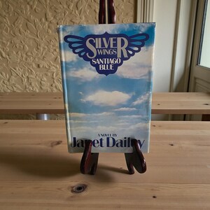 May include: A hardcover book titled "Silver Wings Santiago Blue" by Janet Dailey. The cover features a blue sky with clouds and the title in a stylized font. The book is displayed on a small wooden easel.