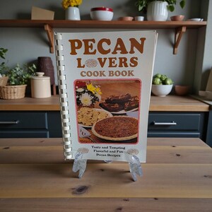 Op de afbeelding: Een vintage kookboek getiteld "Pecan Lovers Cook Book" met een spiraalbinding. De omslag toont de titel in bruine letters en een afbeelding van pecannoten desserts. Het kookboek staat op een transparante plastic standaard.