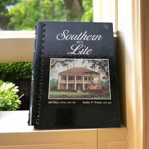 Könnte beinhalten: Ein schwarzes spiralgebundenes Kochbuch mit dem Titel "Southern But Lite" mit einem Foto eines weißen Hauses mit einer Veranda auf dem Einband. Der Text "Jen Bays Avis, LDN, RD" und "Kathy F. Ward, LDN, RD" ist unter dem Foto gedruckt.