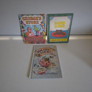 May include: Three vintage children's books are displayed. The covers feature colorful illustrations and titles: "George's Store," "Sand Cake," and "Pirates Ahoy!" The books are arranged on a light surface, showcasing their unique artwork and design.