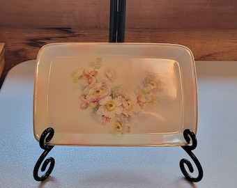 May include: A white ceramic serving tray with a floral design in shades of pink, yellow, and green. The tray is on a black metal stand with two curled legs.