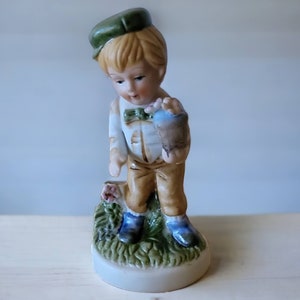 Può includere: Una statuetta in porcellana di un ragazzo con un cappello verde e pantaloni marroni, che tiene un piccolo uccello nella mano. La statuetta è in piedi su una base verde con fiori.