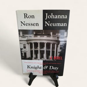 Peut inclure: Un livre intitulé "Knight & Day" de Ron Nessen et Johanna Neuman. La couverture présente une image en noir et blanc d'un bâtiment avec des colonnes. Le livre est soutenu par un support noir. Il s'agit d'un roman.