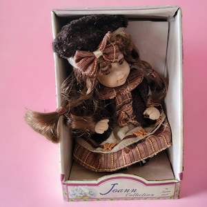 May include: A porcelain doll with brown hair and a brown hat with a brown and white bow. The doll is wearing a brown and gold dress with a plaid pattern. The doll is in a box with the words "Joann Collection" on the front.