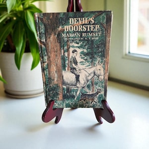 Puede incluir: Libro antiguo "Devil's Doorstep" de Marian Rumsey, ilustrado por W. T. Mars. La portada muestra a un niño montado en un burro en un bosque. Se exhibe en un caballete de madera rojo oscuro.