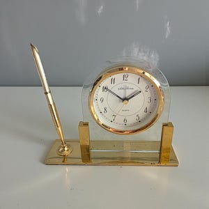 Peut inclure: Une horloge de bureau dorée avec un porte-stylo assorti. L'horloge a un cadran clair et arqué avec une lunette dorée et un cadran blanc. Le cadran de l'horloge a des chiffres et des aiguilles noirs. Le stylo est doré et se tient droit.
