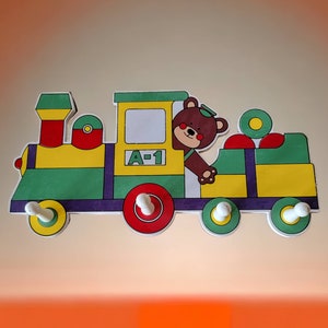 May include: A wooden toy train with a bear conductor. The train is yellow, green, and purple with red wheels. The bear is brown with a yellow hat and is holding a steering wheel. The train has the number A-1 on the side.