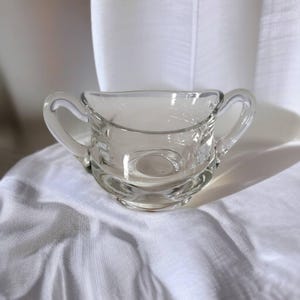 May include: A clear glass sugar bowl with two handles. The bowl has a rounded shape with a decorative base and is sitting on a white cloth. The sugar bowl is empty and appears to be vintage.