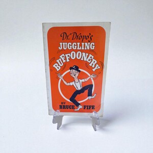 Peut inclure: Couverture de livre vintage pour "Dr. Dropo's Juggling Buffoonery" de Bruce Fife. La couverture présente un fond orange avec une illustration de dessin animé d'un homme jonglant. Le titre et le nom de l'auteur sont en texte blanc et noir.