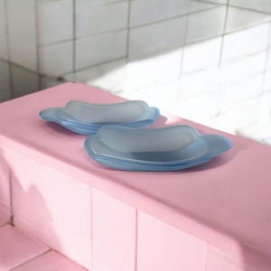 Puede incluir: Dos jaboneras de vidrio azul claro con forma de nube se exhiben sobre una superficie rosa. Los platos tienen un aspecto esmerilado y están uno al lado del otro. El fondo presenta azulejos blancos y luz natural. Miden unos 15 cm.