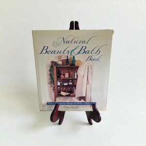 May include: A vintage book titled "The Natural Beauty & Bath Book" displayed on a small wooden easel. The book cover features an illustration of a wooden cabinet filled with bath products and a hanging towel. The title is in elegant script.