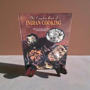May include: A cookbook titled "The Complete Book of Indian Cooking" is displayed on a wooden stand. The cover features images of various Indian dishes. The book is edited by Veronica Sperling & Christine McFadden.