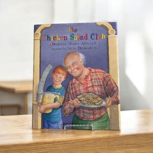 Puede incluir: Un libro infantil titulado "The Chicken Salad Club" se muestra sobre una superficie de madera. La portada presenta una ilustración de un anciano y un niño. El hombre sostiene un plato de sándwiches y el niño lleva un recipiente.
