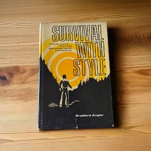May include: A vintage book titled "Survival With Style" with a black cover featuring a silhouette of a person and a yellow sun design. The book's title is in large, bold letters. The author's name, Bradford Angier, is also visible.