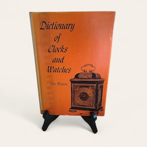 May include: An orange book titled "Dictionary of Clocks and Watches" by Eric Bruton. The cover features a detailed illustration of a clock. The book is propped up on a black stand.