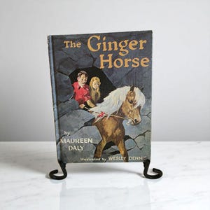 Puede incluir: Libro antiguo titulado "The Ginger Horse" de Maureen Daly, ilustrado por Wesley Dennis. La portada muestra un caballo y dos niños. El libro se exhibe en un soporte de metal negro.