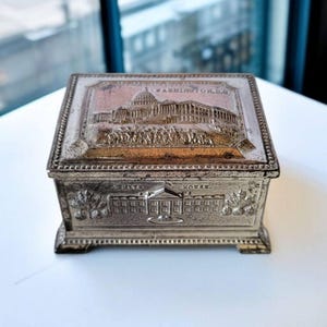 Peut inclure: Une boîte à bijoux en métal argenté avec une image en relief du Capitole des États-Unis et le texte "WASHINGTON, D.C." sur le dessus. Le fond de la boîte présente une image en relief de la Maison-Blanche et le texte "WHITE HOUSE".