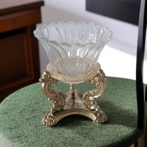 May include: A clear glass bowl with a decorative, ornate gold metal stand. The bowl has a fluted design and the stand has a round base with three legs.