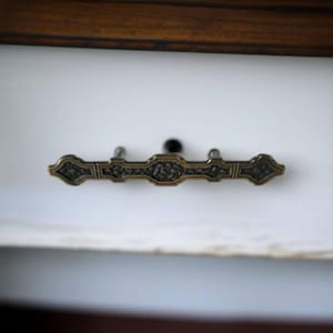 May include: A brass drawer pull with an ornate design. The pull is mounted on a white surface.