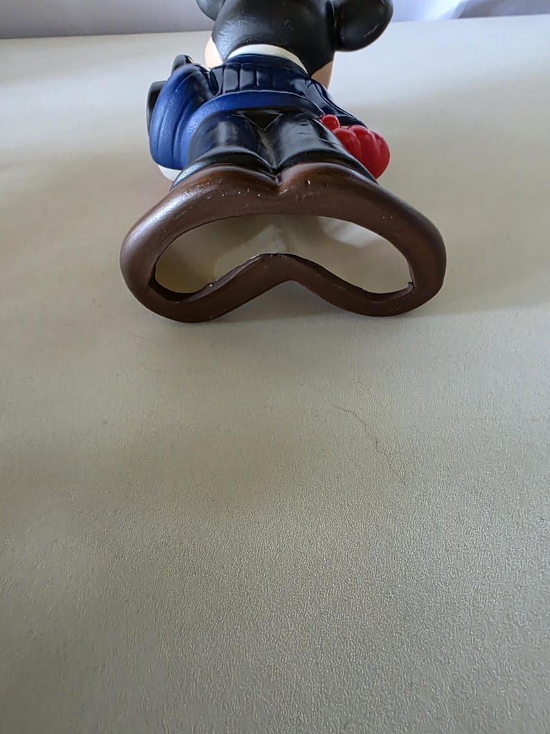 K&ouml;nnte beinhalten: Eine Flaschen&ouml;ffner-Figur von Micky Maus aus Keramik. Die Figur tr&auml;gt eine blaue Jacke, braune Hose und rote Handschuhe. Der Flaschen&ouml;ffner ist braun und hat die Form von Mickys Shorts.